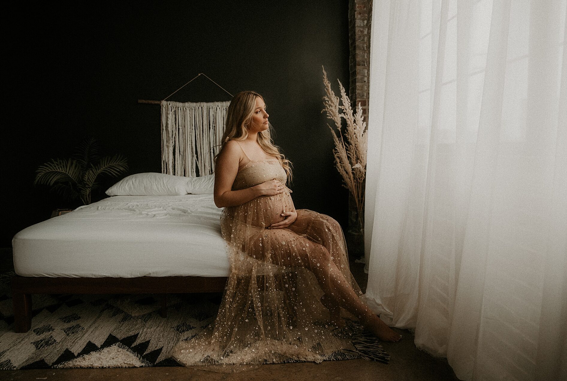 Indoor maternity studio session in St Louis macrame, persian rug, brick and black wall. Expecting mother sitting on bed holding her belly