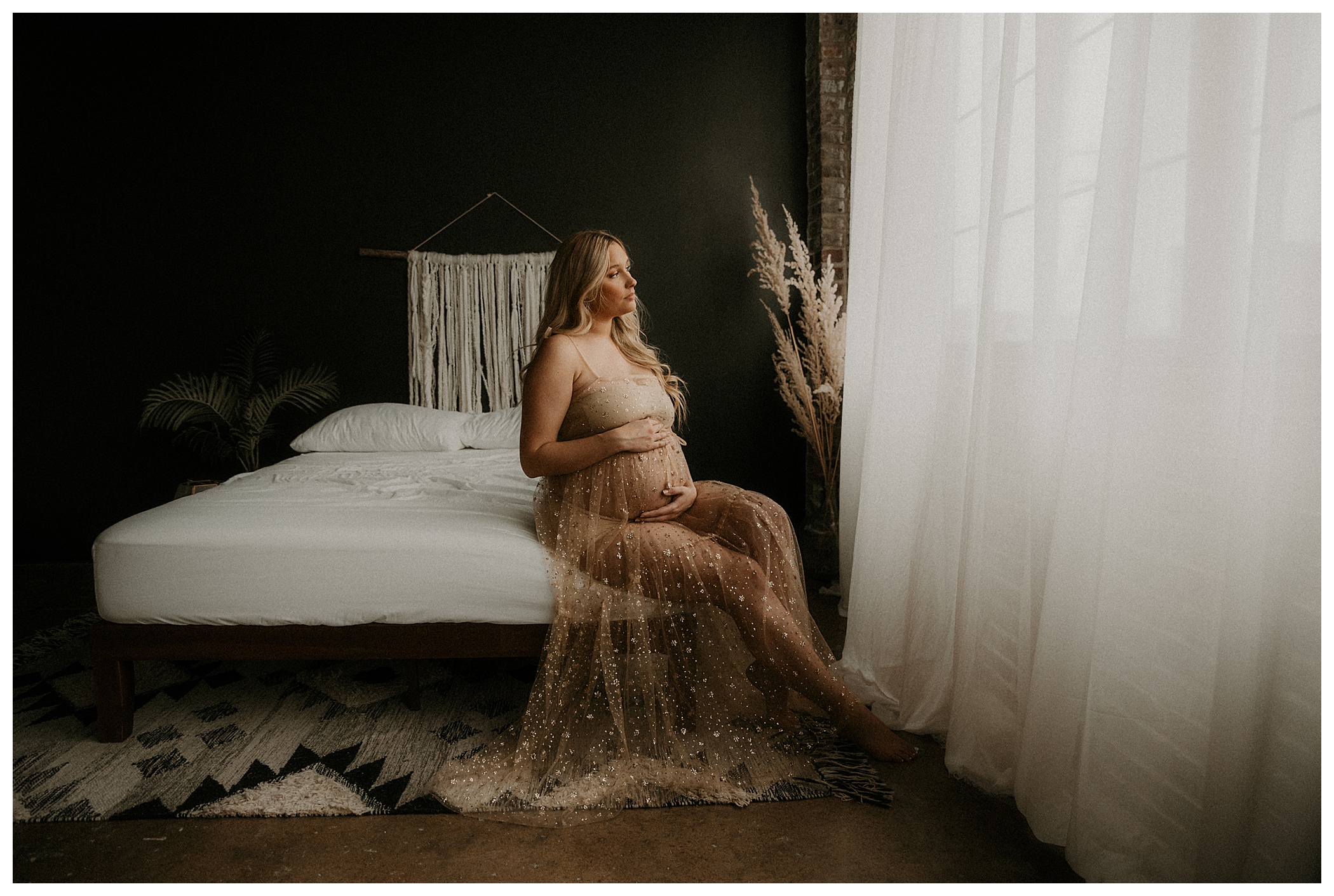 Indoor maternity studio session in St Louis macrame, persian rug, brick and black wall. Expecting mother sitting on bed holding her belly