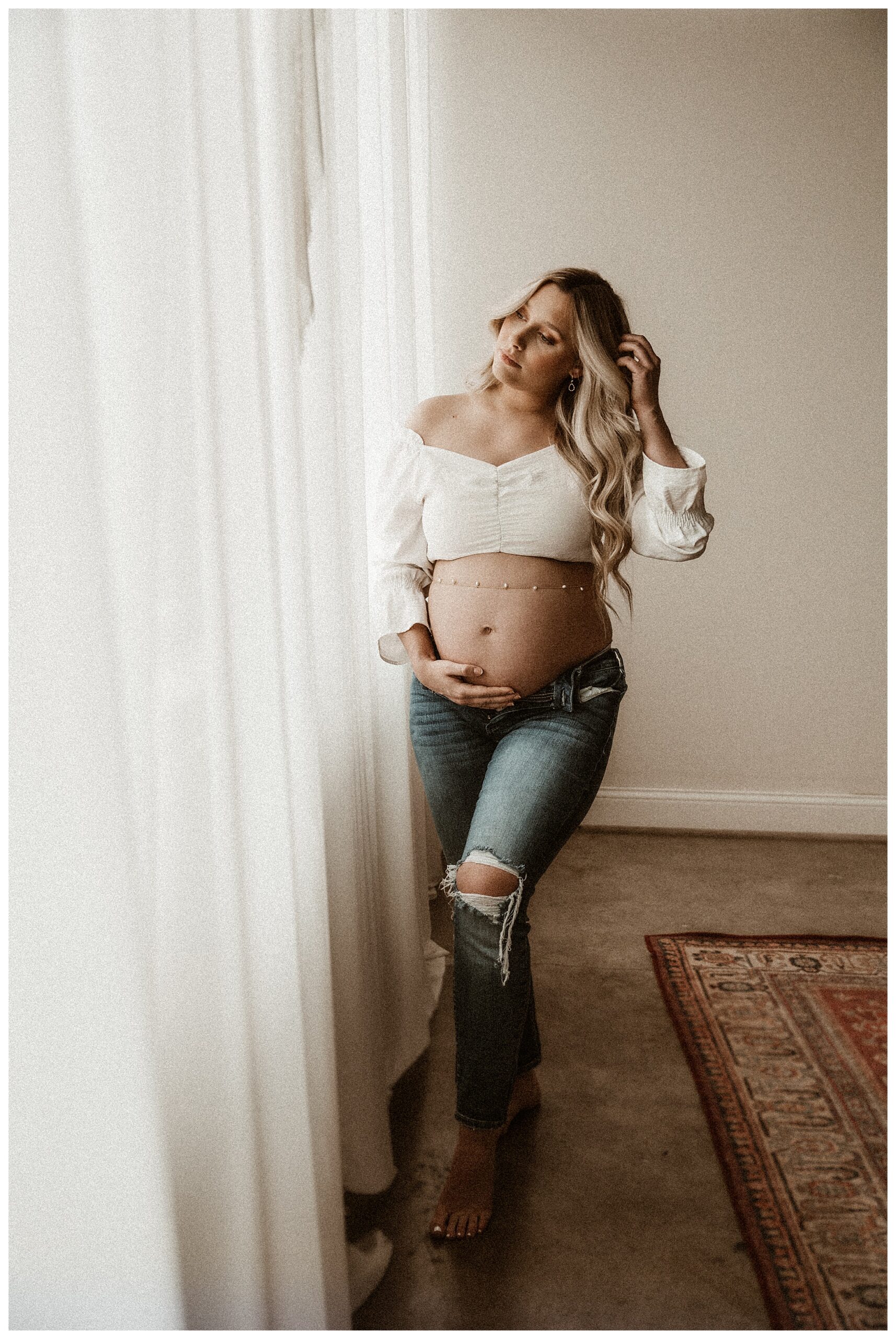 ripped jeans and white shirt maternity session in studio with persian rug. clean and moody portrait of mother fixing her hair