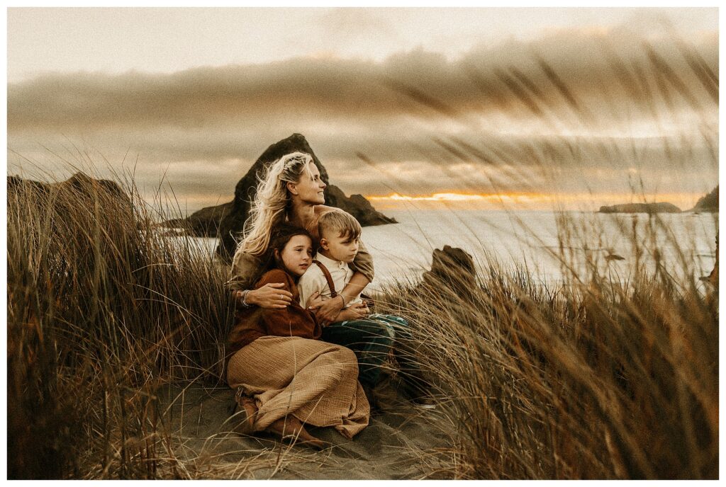 Mother sitting in dunes with tall grass, overlooking the sunset over the ocean in windy romantic outdoor family session at the Oregon coast at Stormy Solis workshop Rebecca Stephenson fine art family photographer St Louis Missouri