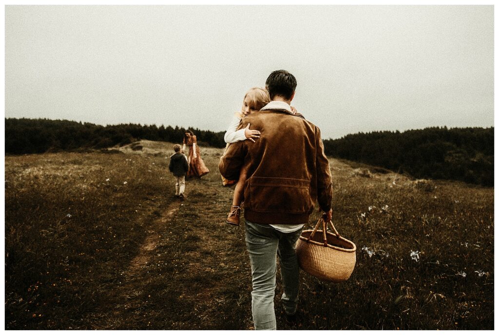 dad walking away from camera while holding a picnic basket, mom playing with her son in background