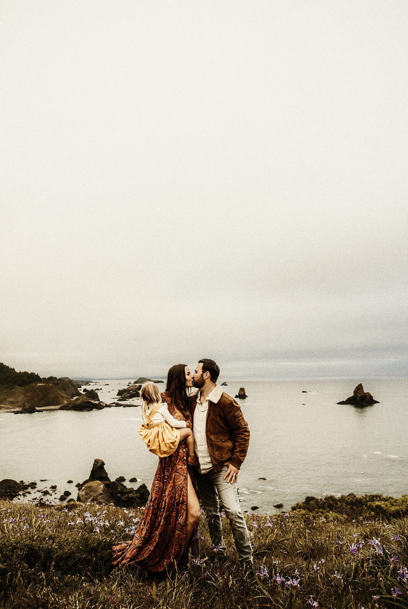 mom and dad kissing while holding their daughter in moody grainy outdoor session by the ocean
